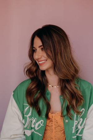 Modeportret van een model in een groen-witte varsityjack met een oranje top tegen een roze achtergrond.
