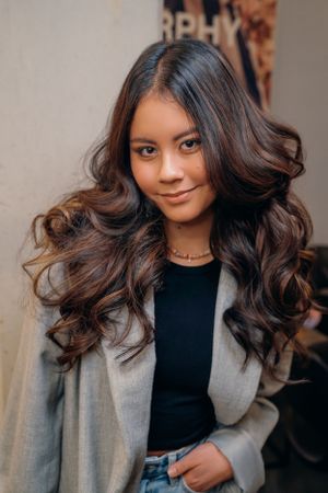 Portret van een vrouw in een zwarte top en grijze blazer met lang, golvend bruin haar, glimlachend naar de camera.