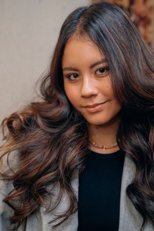 Portret met lang, donker, golvend haar gestyled in zachte krullen, gekleed in een zwarte top en een grijze blazer.