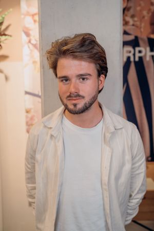 Een persoon gekleed in een witte jas en T-shirt staat tegen een muur in een binnenruimte.