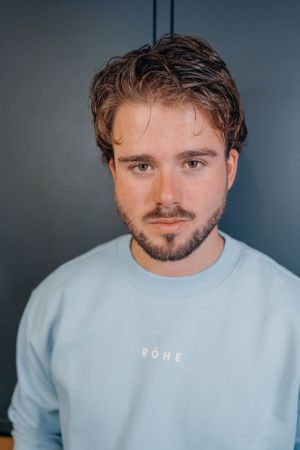 Portretfoto tegen een donkere achtergrond van een man met golvend bruin haar en gezichtsbeharing, gekleed in een lichtblauwe sweatshirt.