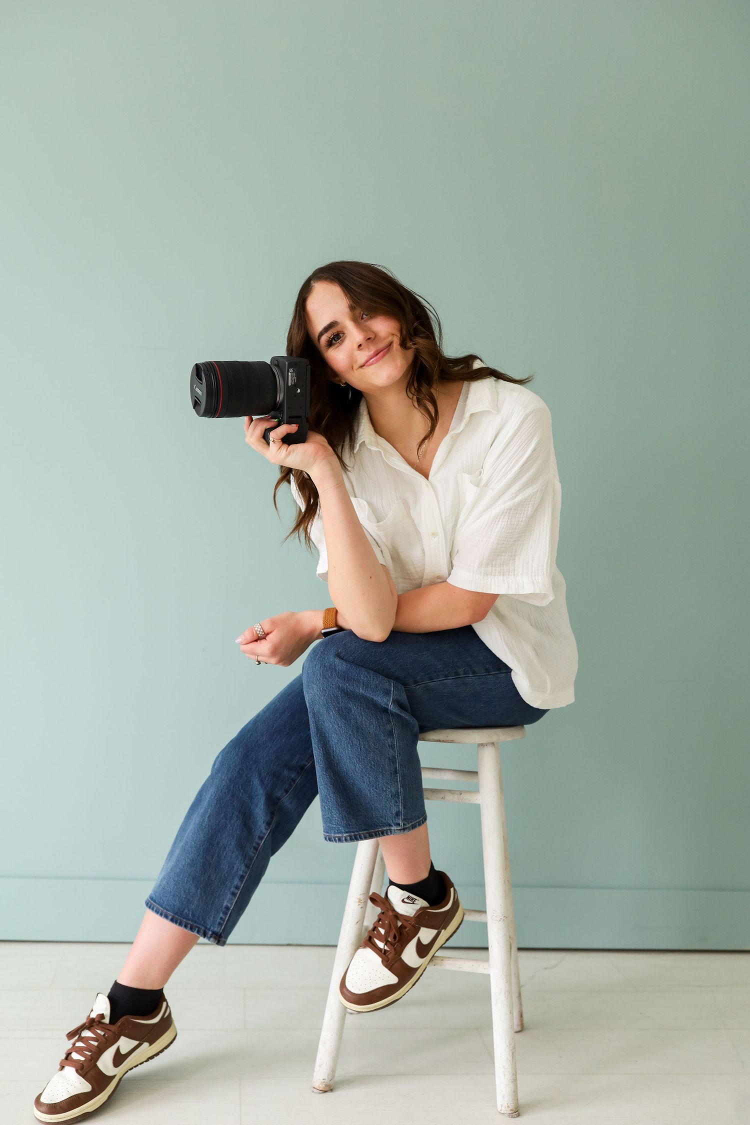 Female photographer sits on a stool holding a DSLR camera, smiling against a soft mint green background.