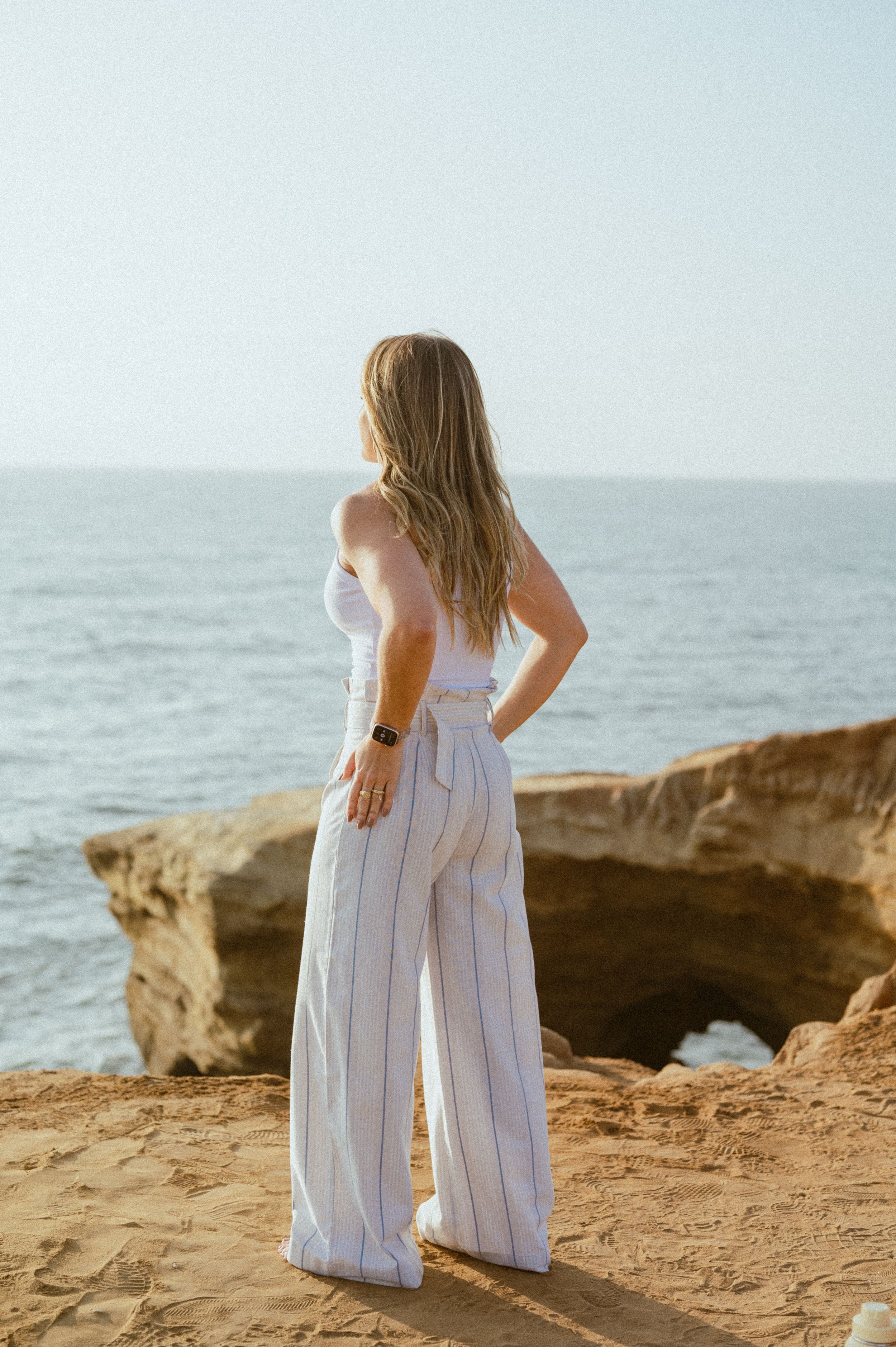 Woman in flowing white dress poses on rocky coastal cliff during daytime.