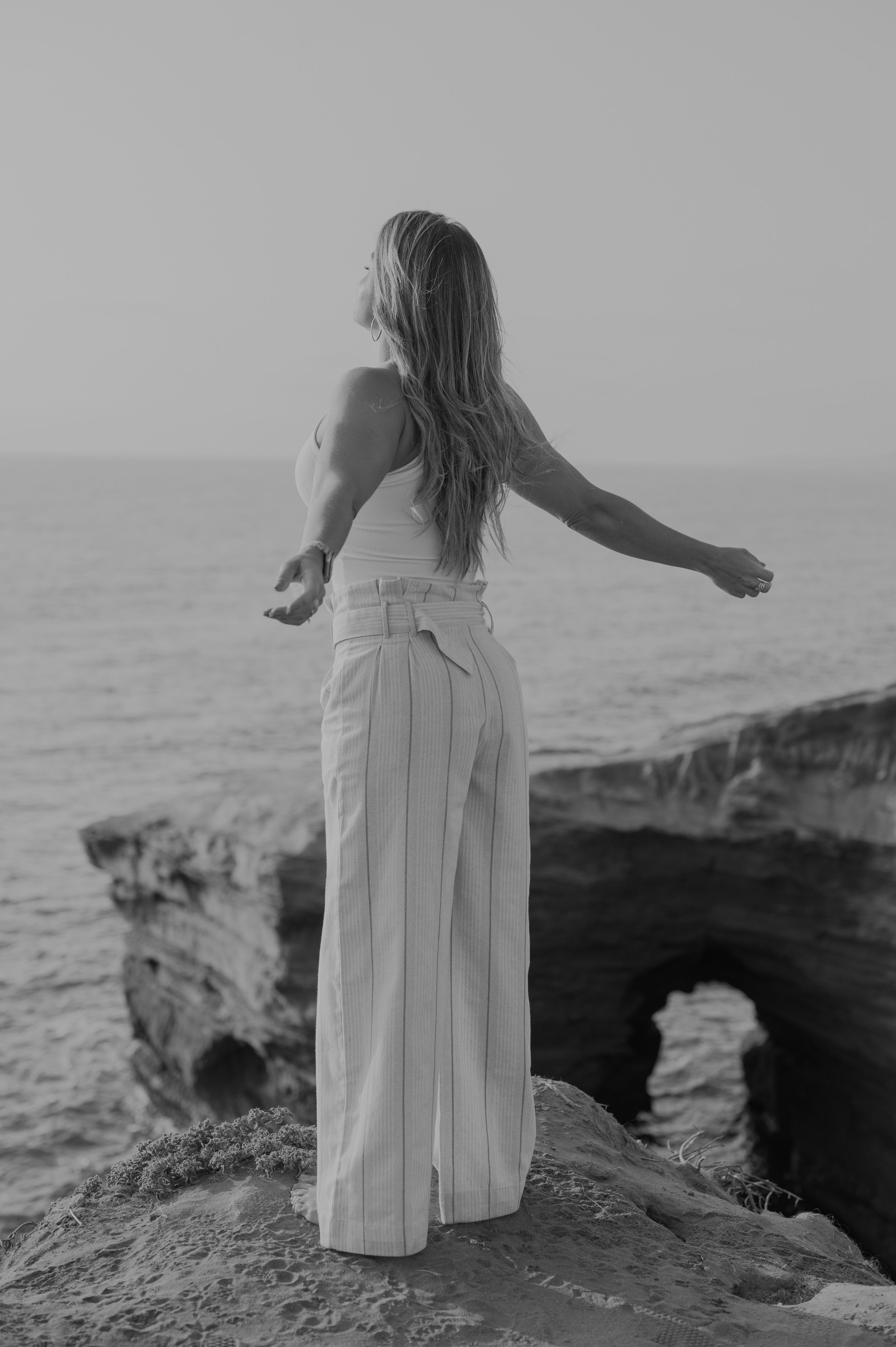 Person in white dress standing on coastal cliff edge overlooking ocean.