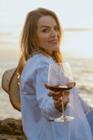 Person enjoying a glass of red wine at sunset on a beach.