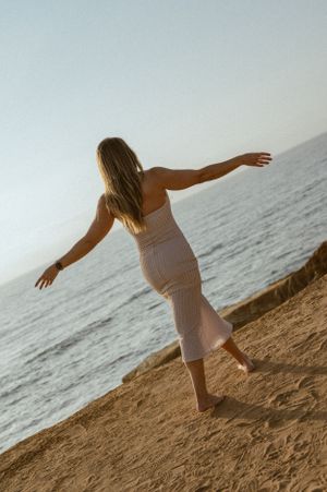 Figure in white dress balancing with arms outstretched on coastal rocks at sunset.