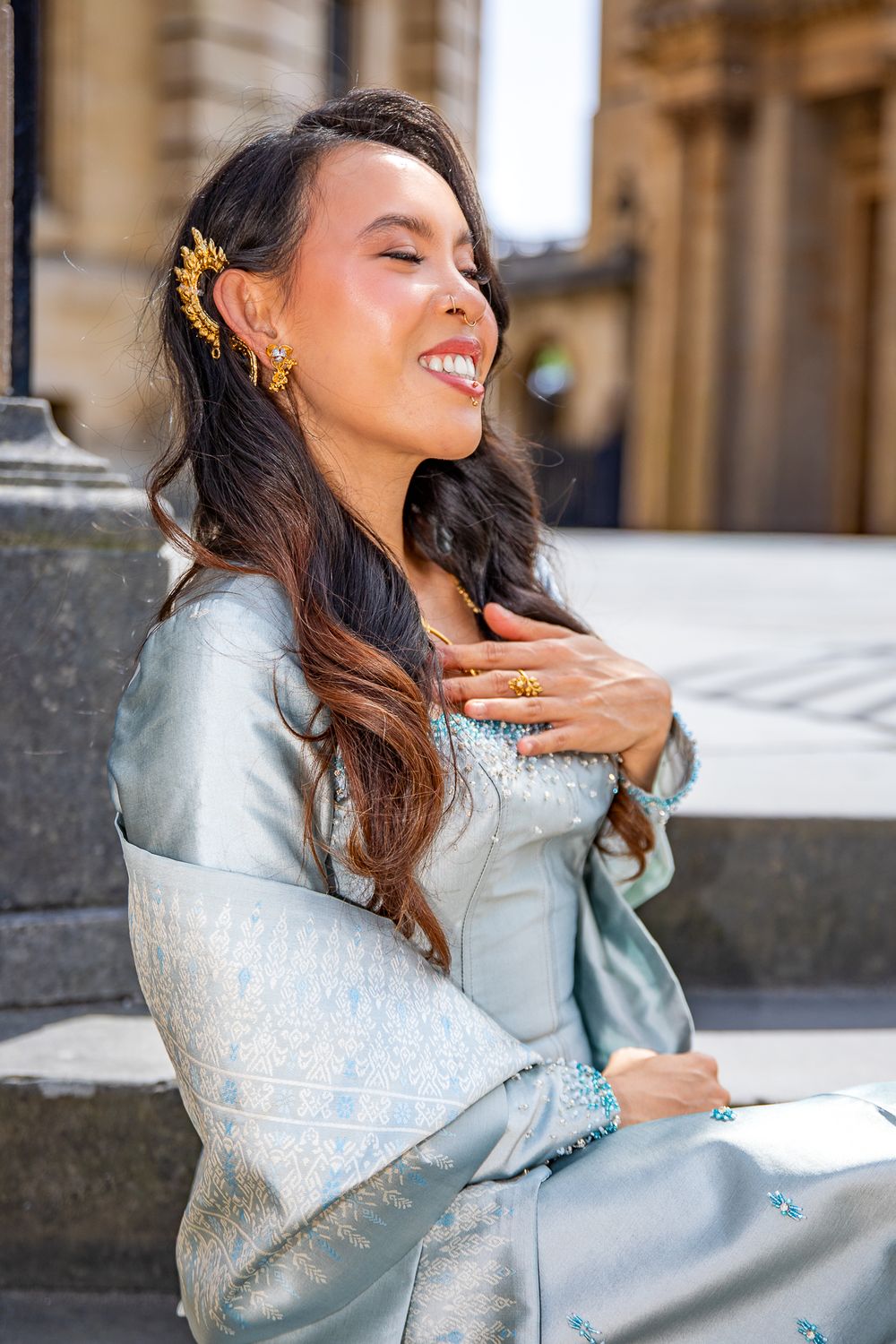 Smiling woman in light blue traditional dress with gold hair accessories and jewelry, sitting outdoors.