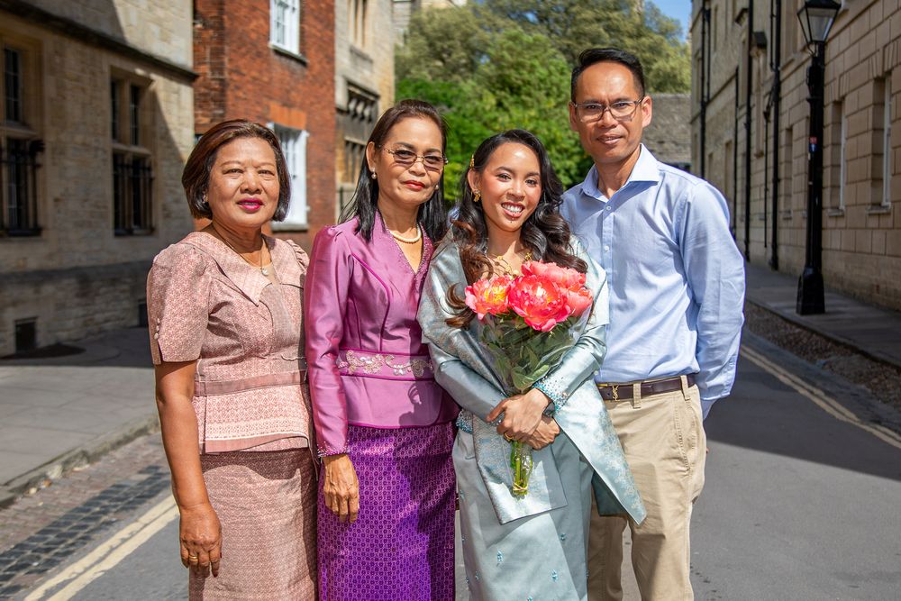 Family of four posing together on a sunny cobblestone street, woman holding pink flowers at graduation celebration.