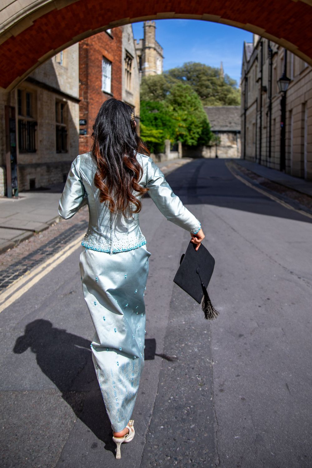 Woman in light blue traditional dress holding graduation cap, walking under stone arch on sunny day.