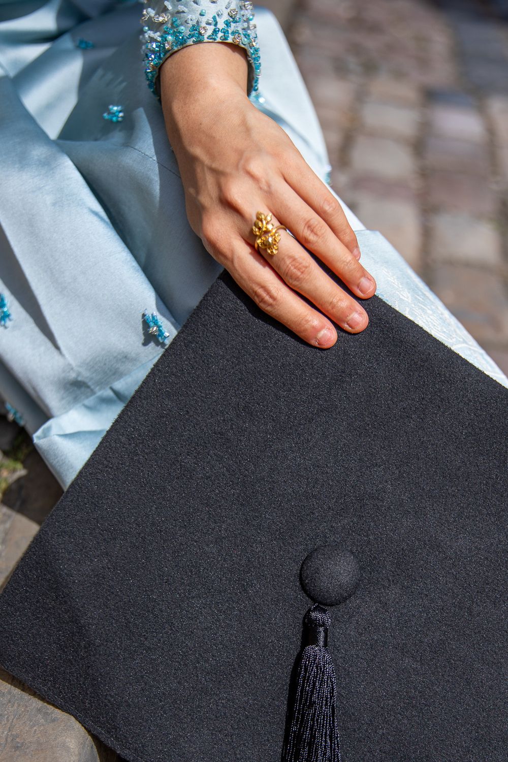 A graduate in a light blue gown holds a black mortarboard cap, wearing a gold ring, on a cobblestone background.