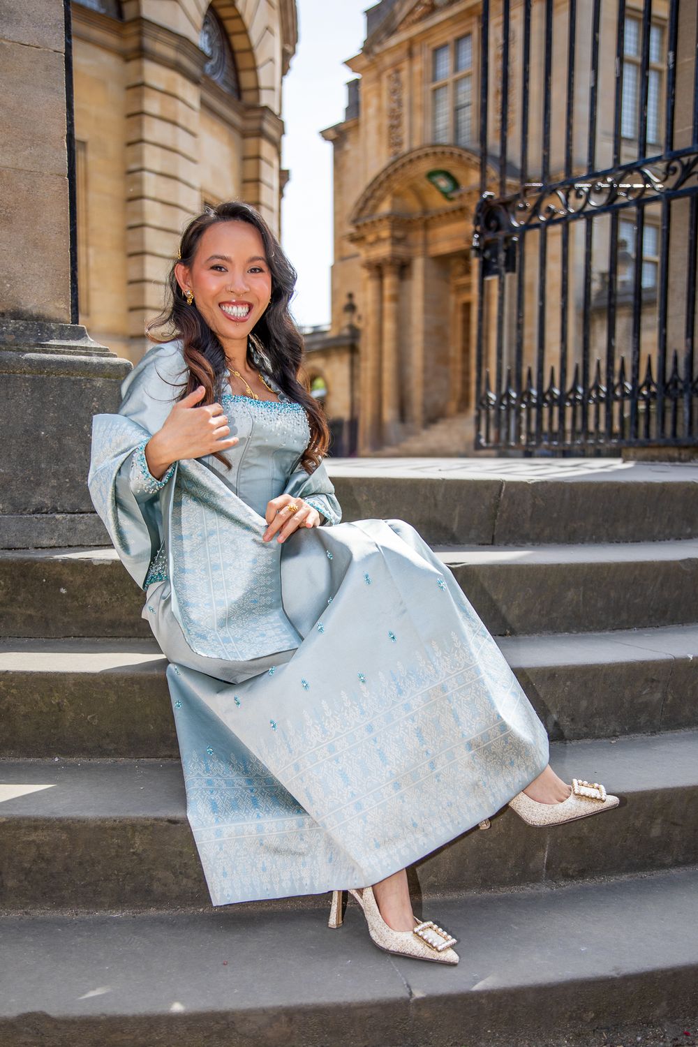 Woman in light blue gown sitting on stone steps of a classical building, smiling elegantly in heels.