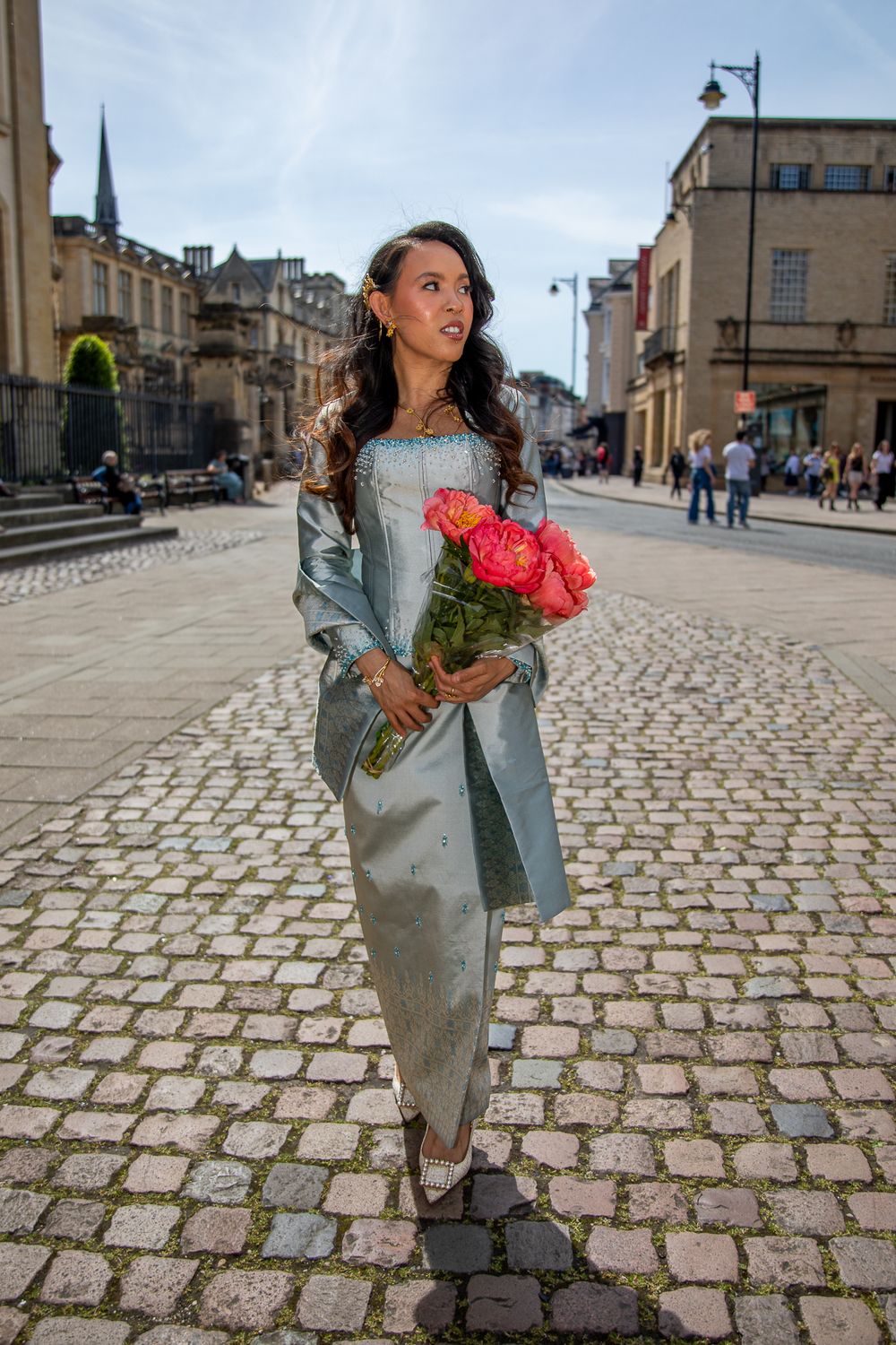 Woman in blue gown holding pink roses on cobblestone street in historic European city.