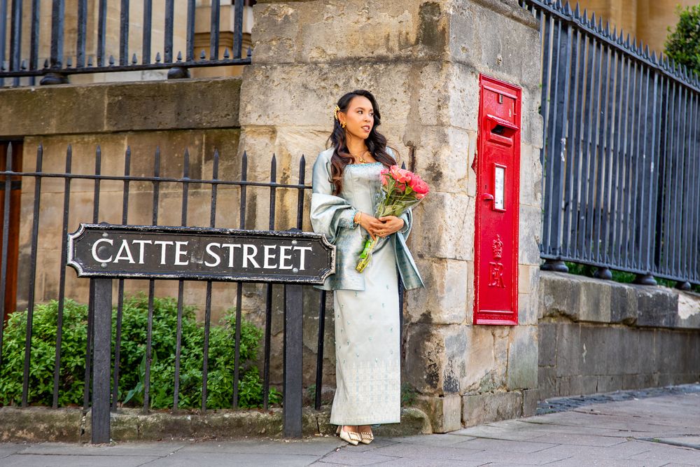 Woman in elegant dress holding flowers by Catte Street sign and red postbox in Oxford.