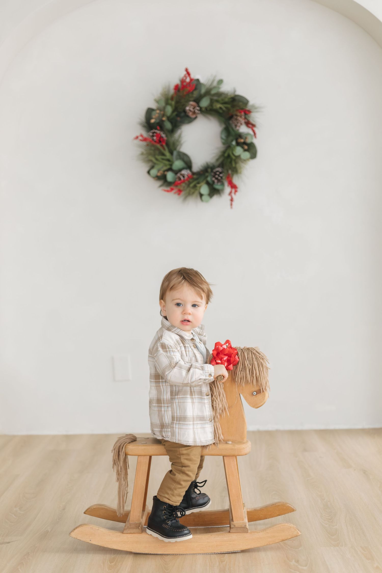 Toddler in a plaid shirt and black boots rides a wooden rocking horse holding a red bow beneath a holiday wreath on a white wall.