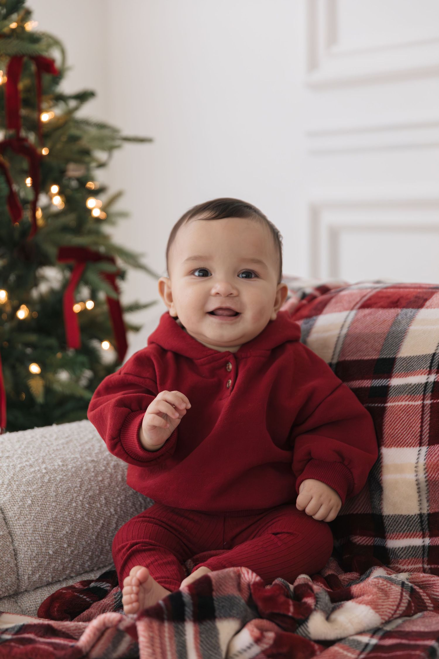 Smiling baby in a red hooded sweatsuit sits on a plaid blanket beside a decorated Christmas tree with red ribbons and warm lights.