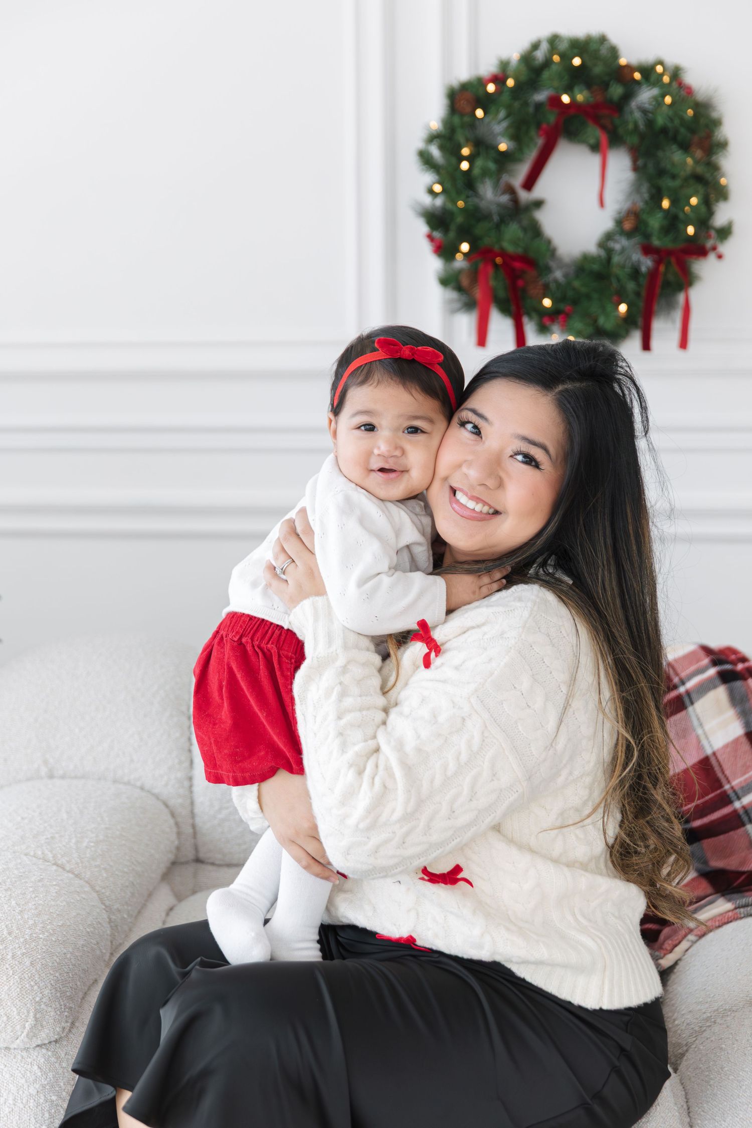 Smiling woman in a cream cable-knit sweater holds a baby in a red skirt and bow near a holiday wreath on a white sofa.