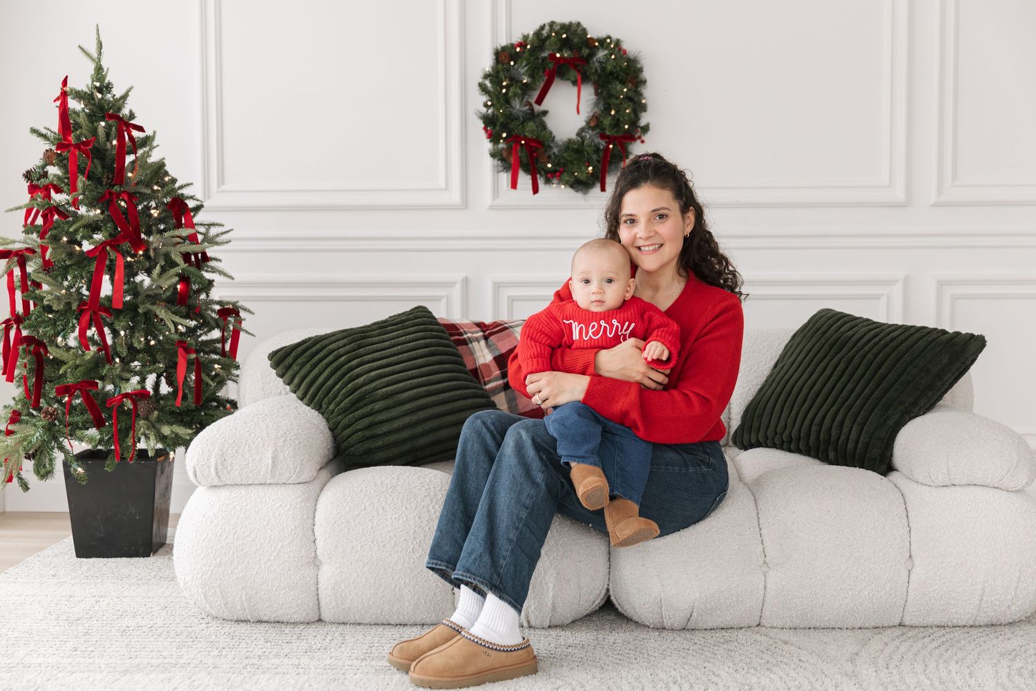 Smiling woman in red sweater holding a baby on a white sofa with green pillows, beside a decorated Christmas tree and holiday wreath.