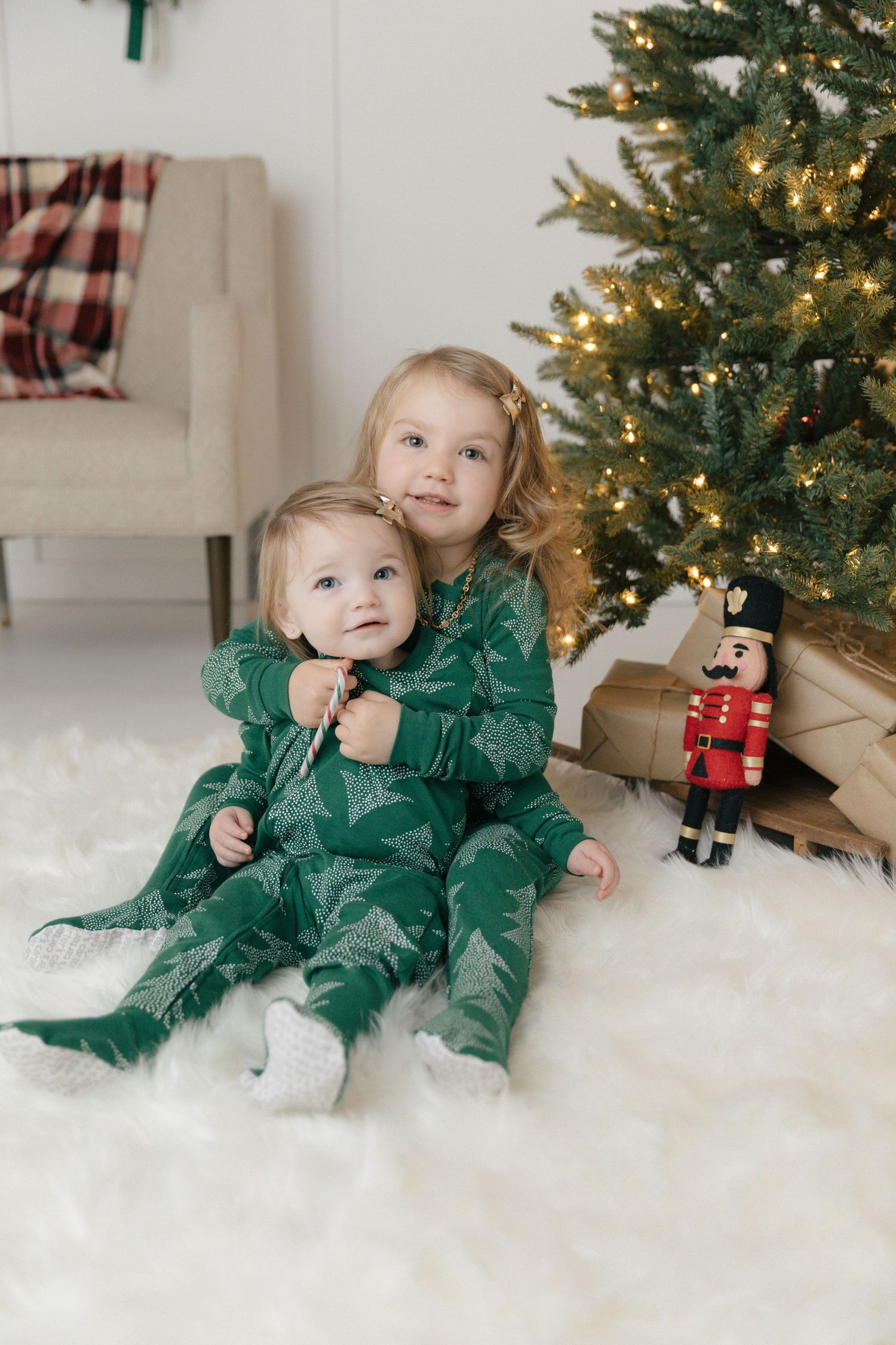Two children in matching green outfits sit on white carpet near a decorated Christmas tree with presents.
