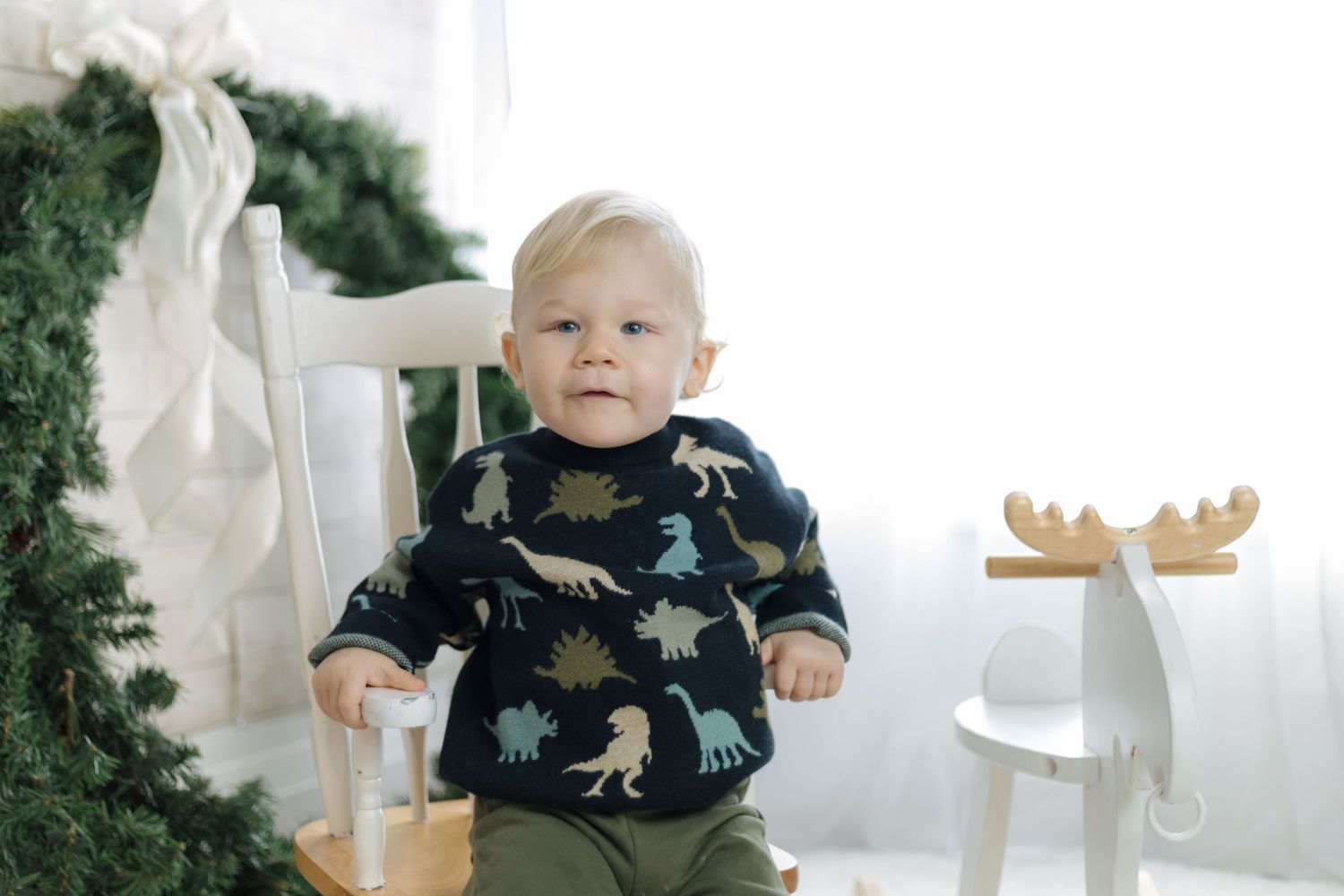 A winter scene with holiday decor featuring a small child sitting in a white chair near greenery.