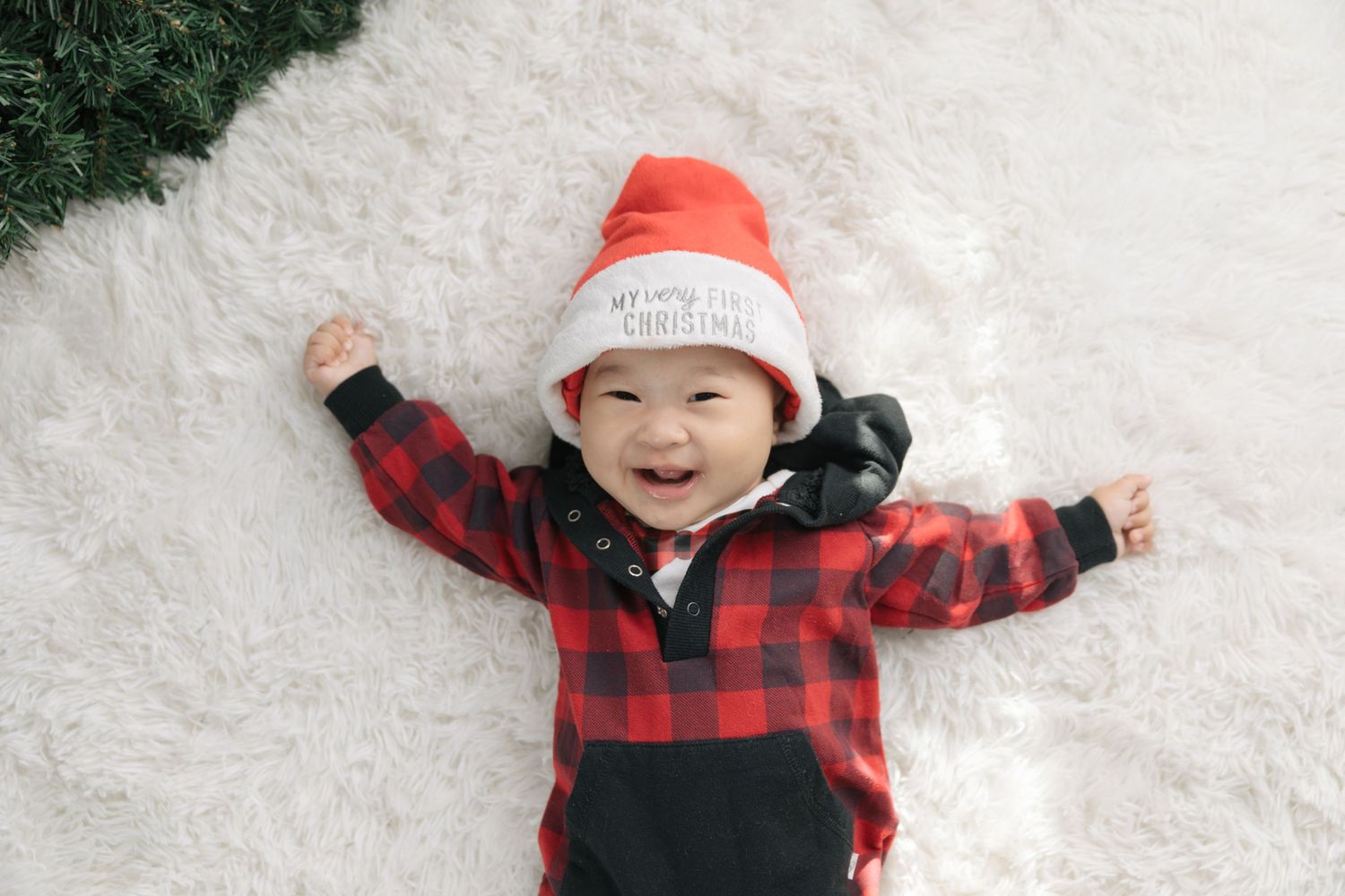 A happy baby in buffalo plaid and santa hat lying on white furry carpet with holiday decorations.