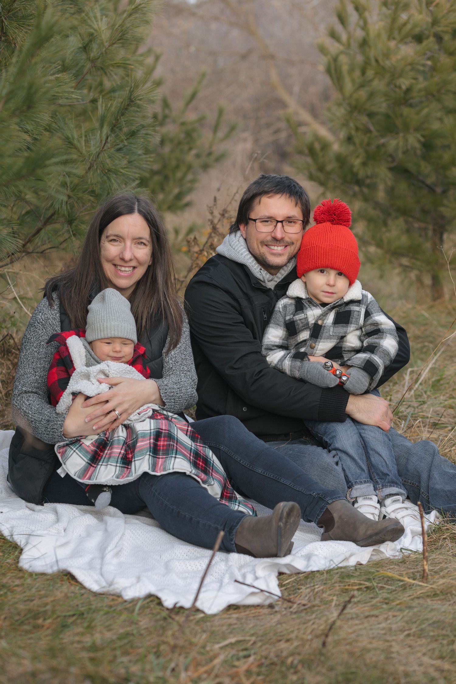 Smiling family of four sits on a white blanket among pine trees, with a baby in a gray hat and a toddler in a red pom-pom hat.