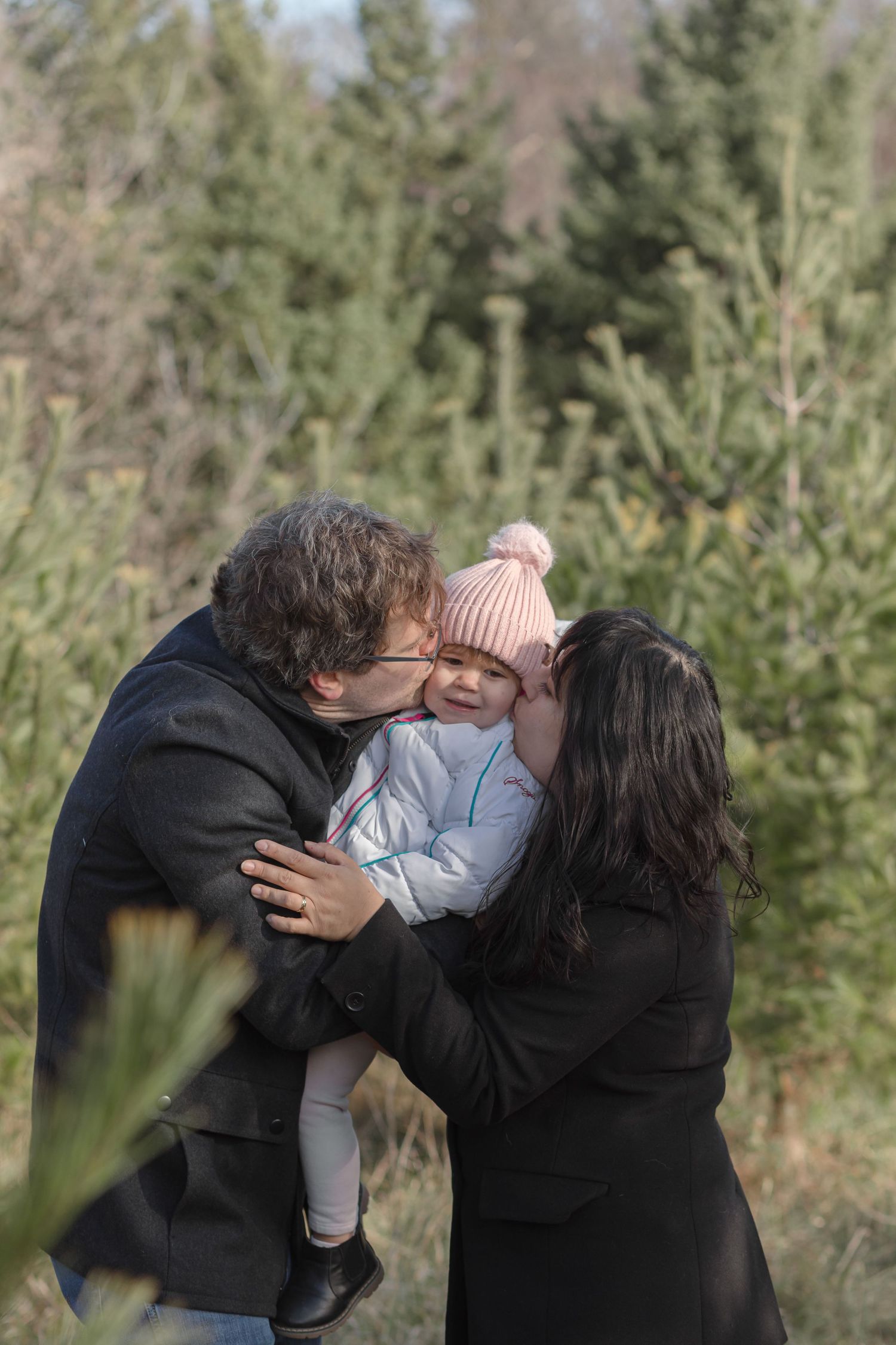 Two parents kiss a smiling toddler in a pink pom-pom hat while standing among evergreen trees at a Christmas tree farm.