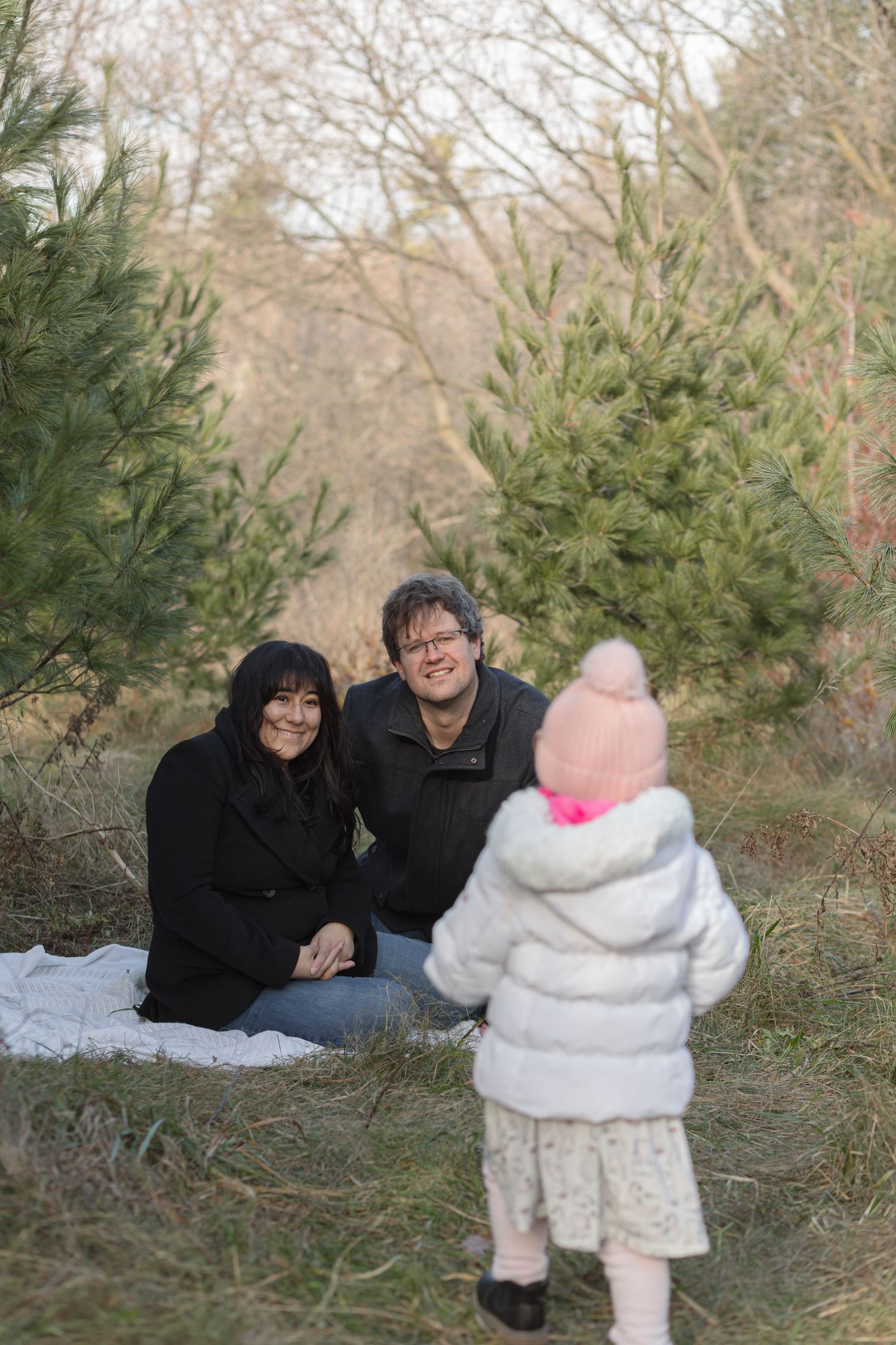 Two smiling adults sit on a white blanket among young pine trees while a toddler in a pink hat and white jacket faces them.