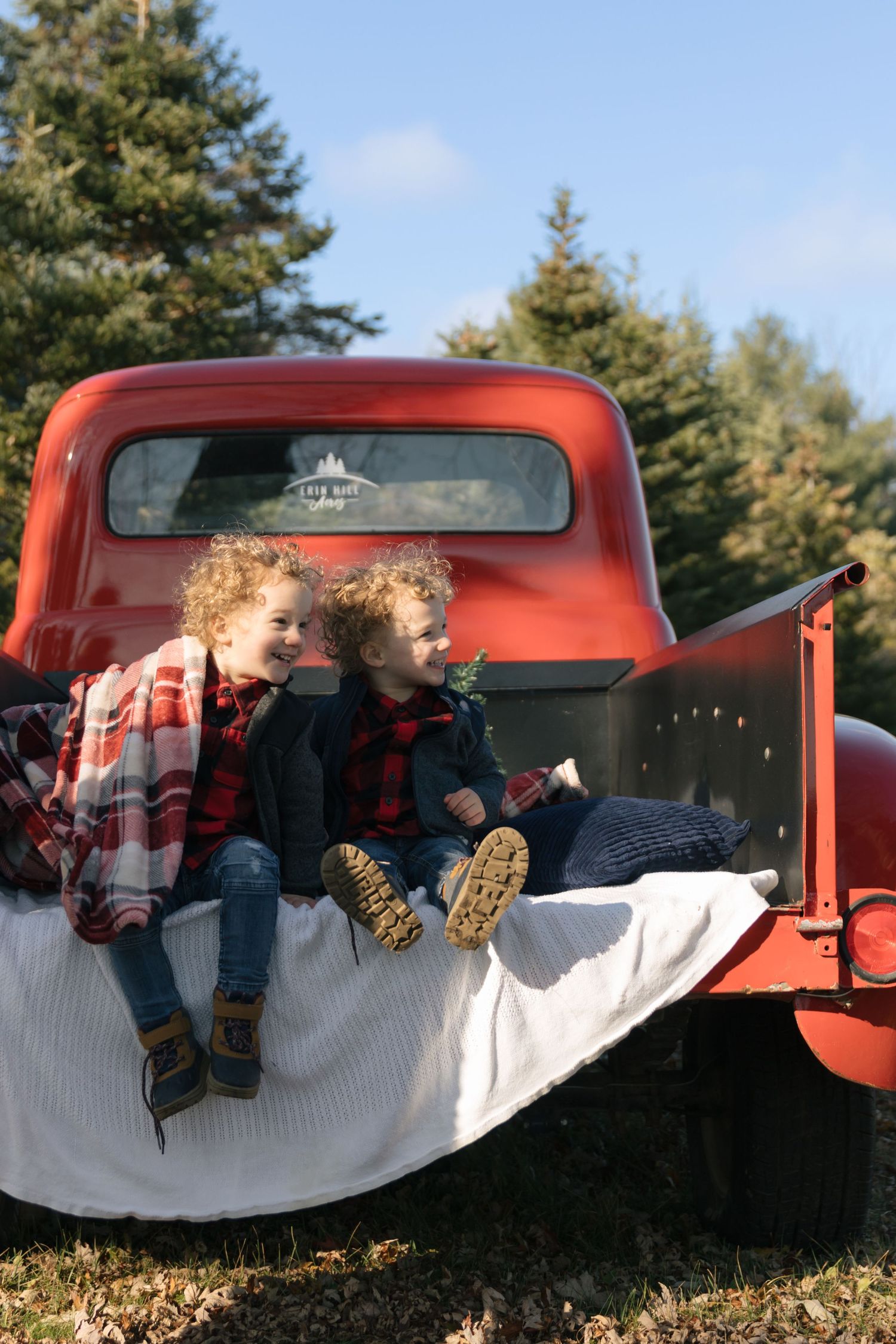 Two children sit in the back of a vintage red pickup truck on a white blanket surrounded by evergreen trees.