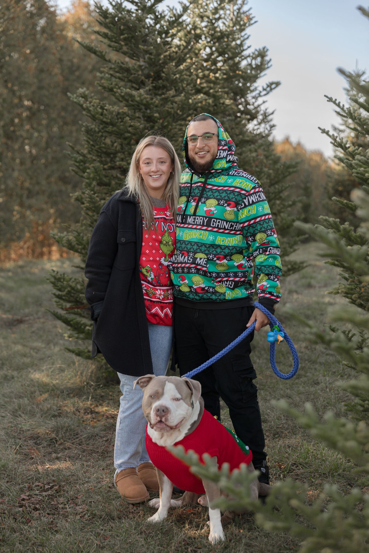 A couple in festive sweaters poses with their dog among evergreen trees at a Christmas tree farm.