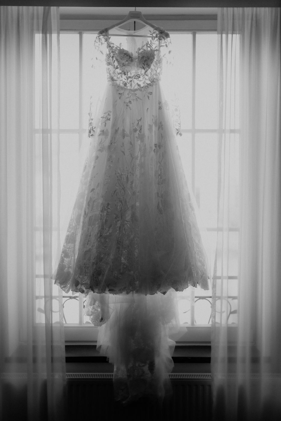 An elegant wedding dress with lace detailing and floral embellishments hangs in a sunlit window creating a dreamy silhouette.
