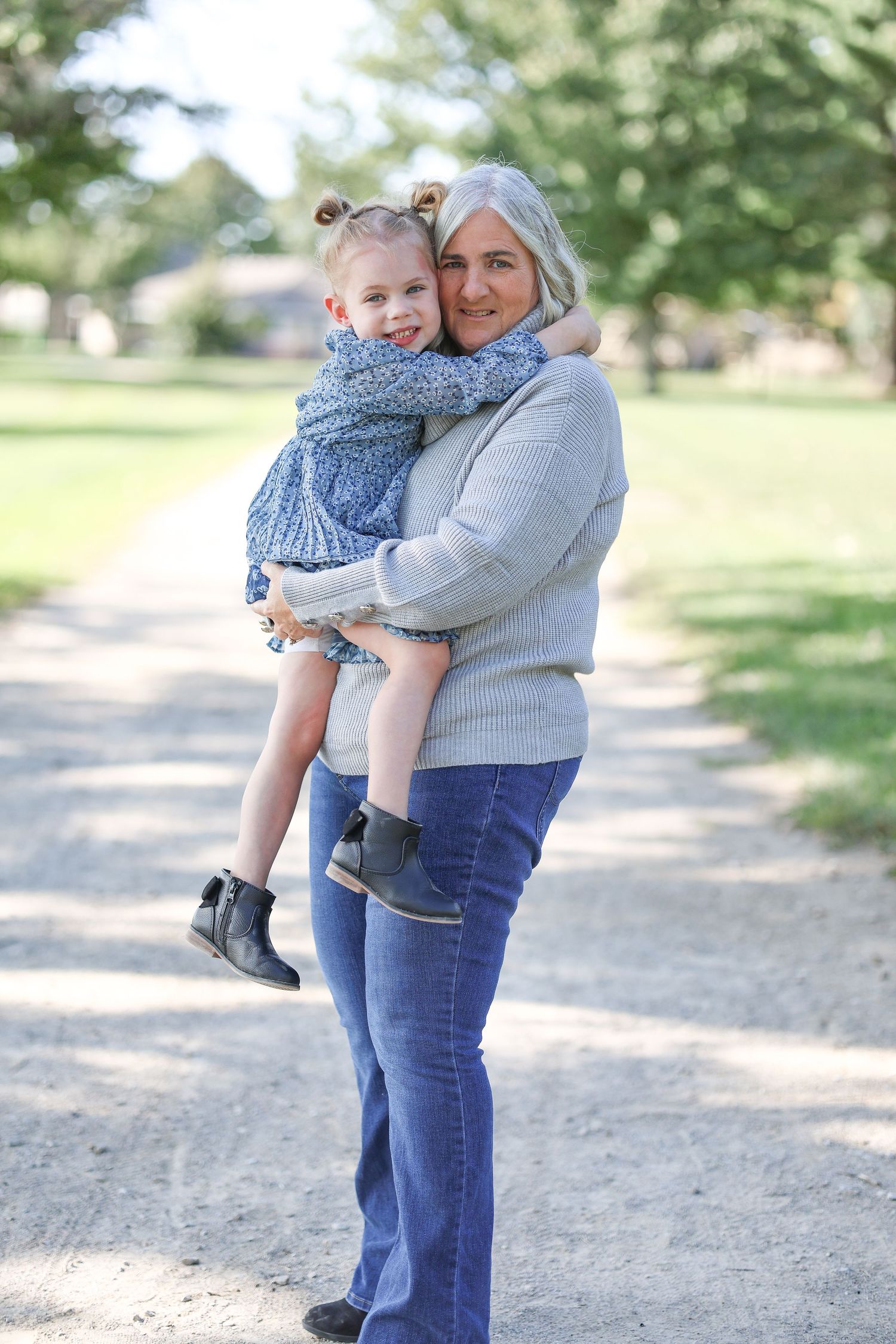 Woman in grey sweater embraces young child wearing blue dress on outdoor path surrounded by greenery.