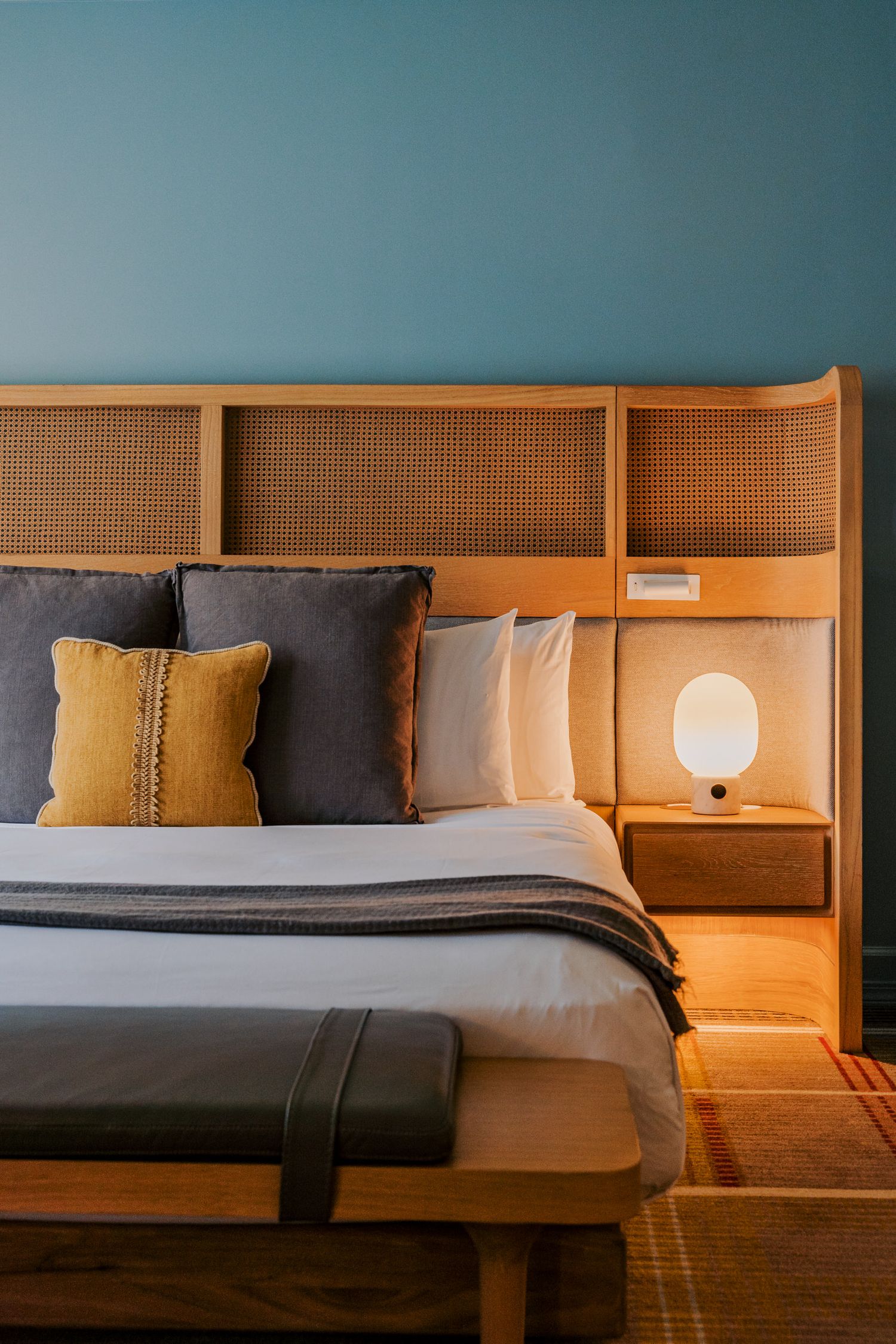 Modern hotel bedroom with wooden headboard, blue accent wall, and mustard throw pillows on a plush bed.