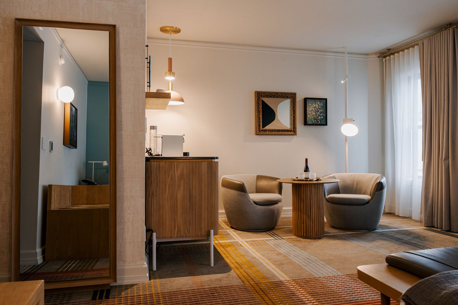 Modern hotel room interior with gray accent chairs, wooden furniture, and contemporary lighting fixtures on a patterned area rug.