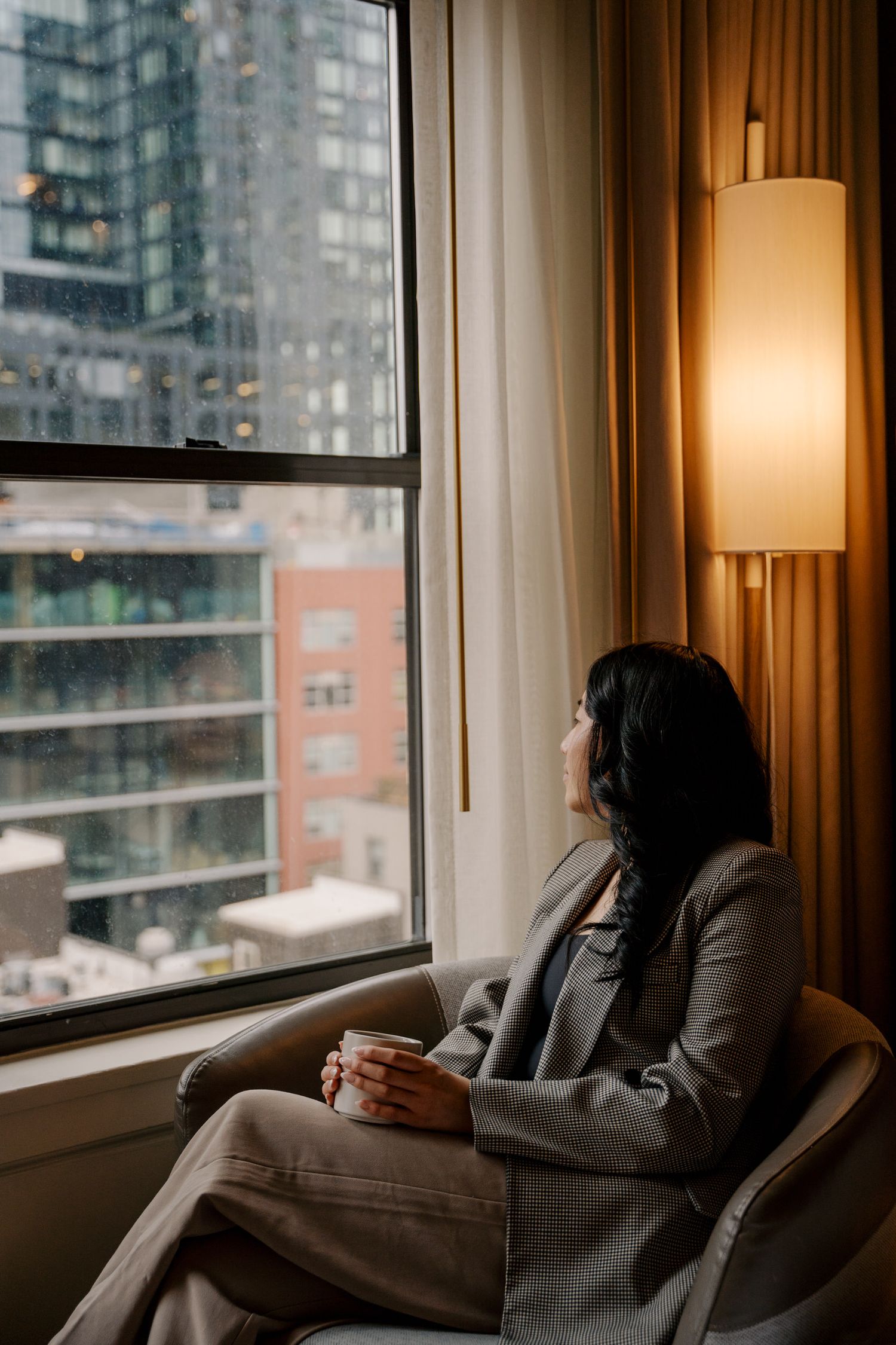 Cozy urban apartment scene with a person relaxing in a chair by the window overlooking city buildings.
