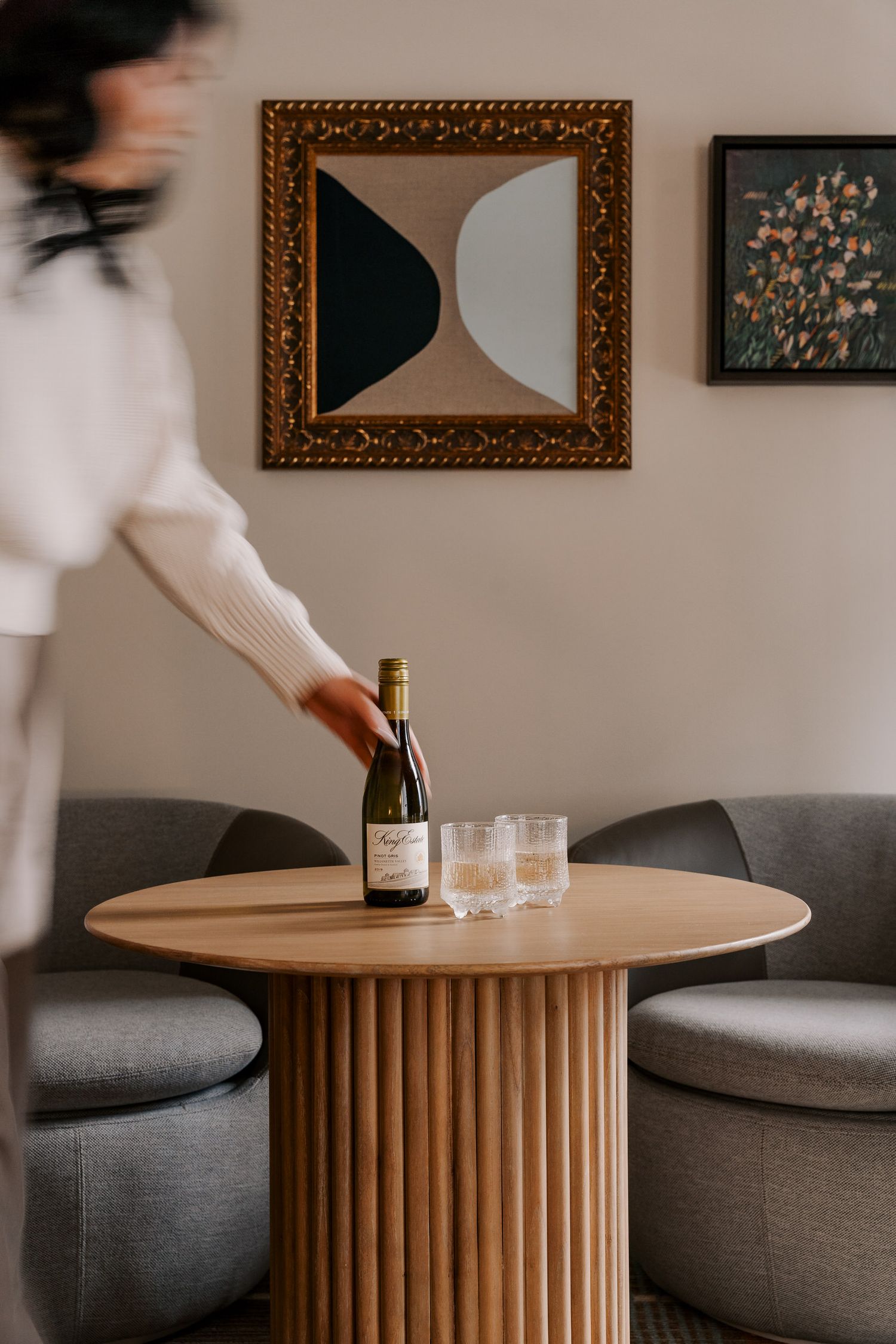 Modern living room with geometric wall art and round wooden table with wine bottle setup.