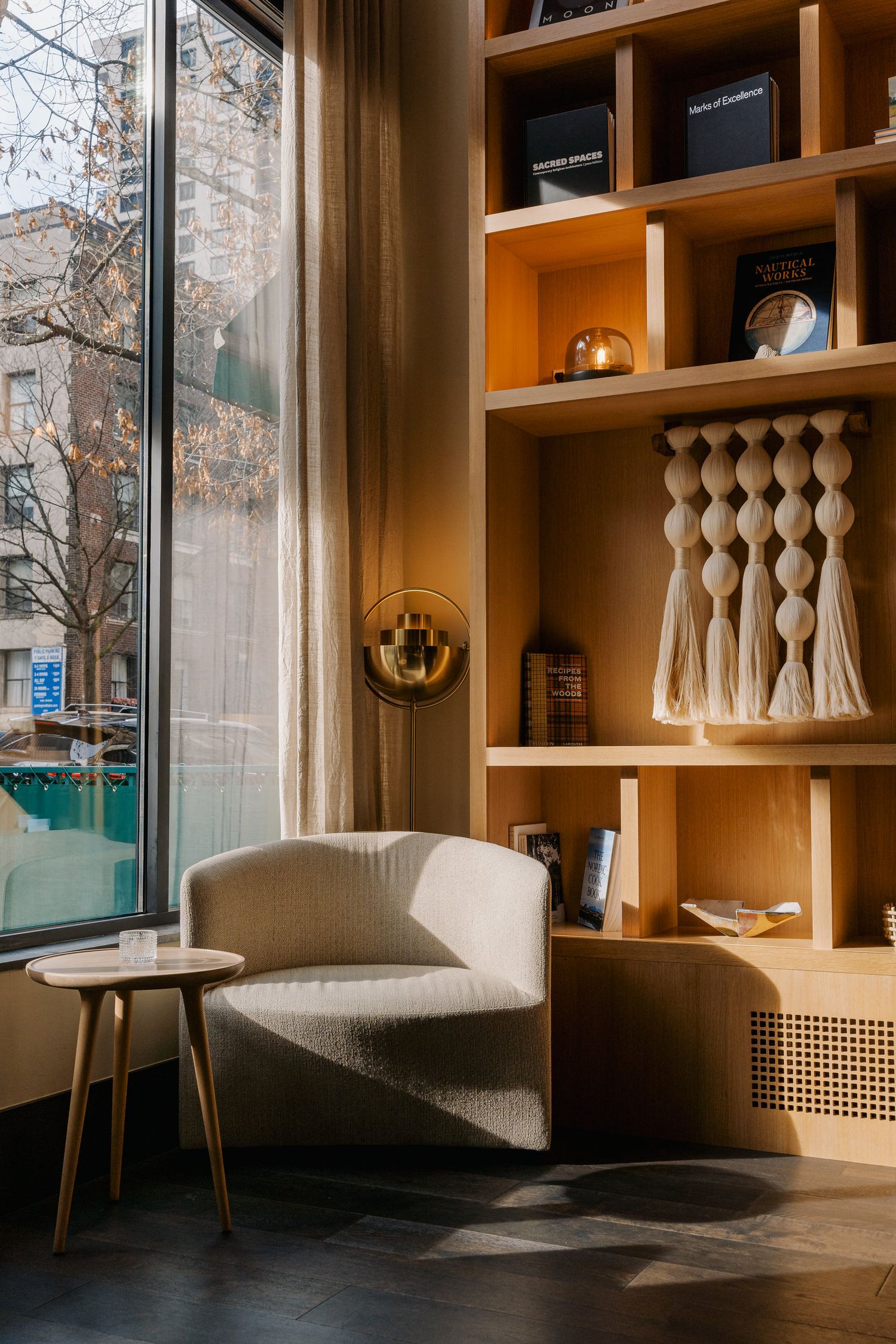 Cozy reading nook with curved cream armchair and wooden built-in shelving displaying decorative objects next to large window.