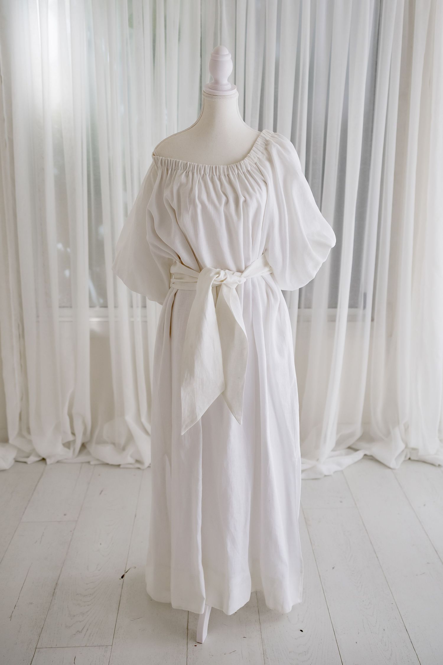 Full-length white off-shoulder peasant dress with tie waist on mannequin.