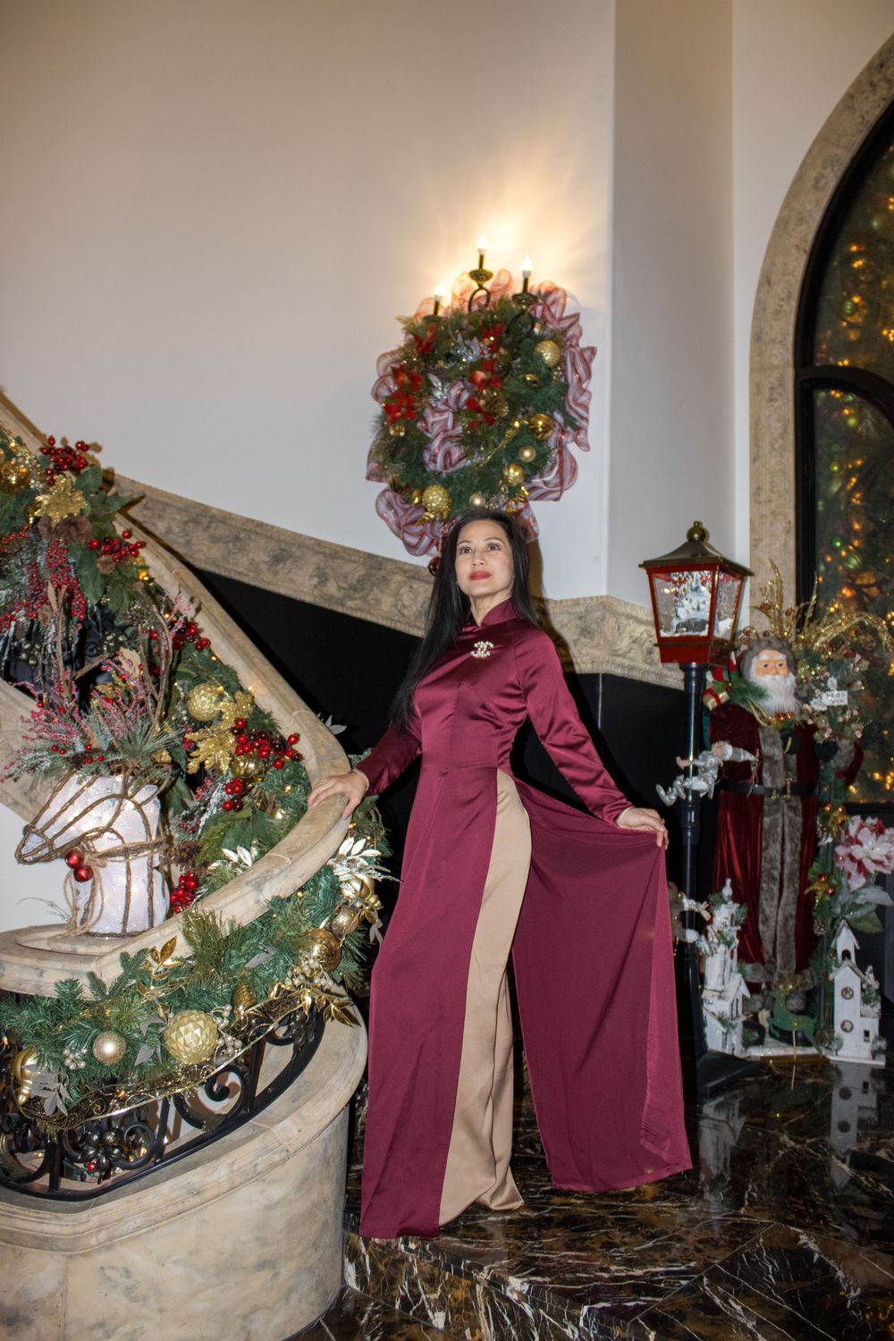 Woman in burgundy and gold ao dai poses on marble staircase surrounded by Christmas decorations.