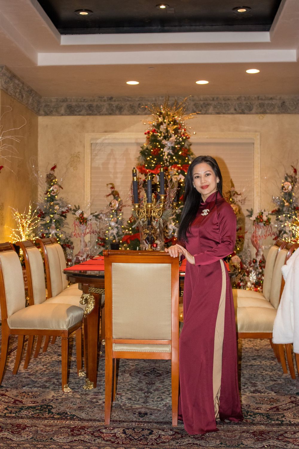 Woman in burgundy ao dai stands in dining room decorated with Christmas tree and holiday decor.
