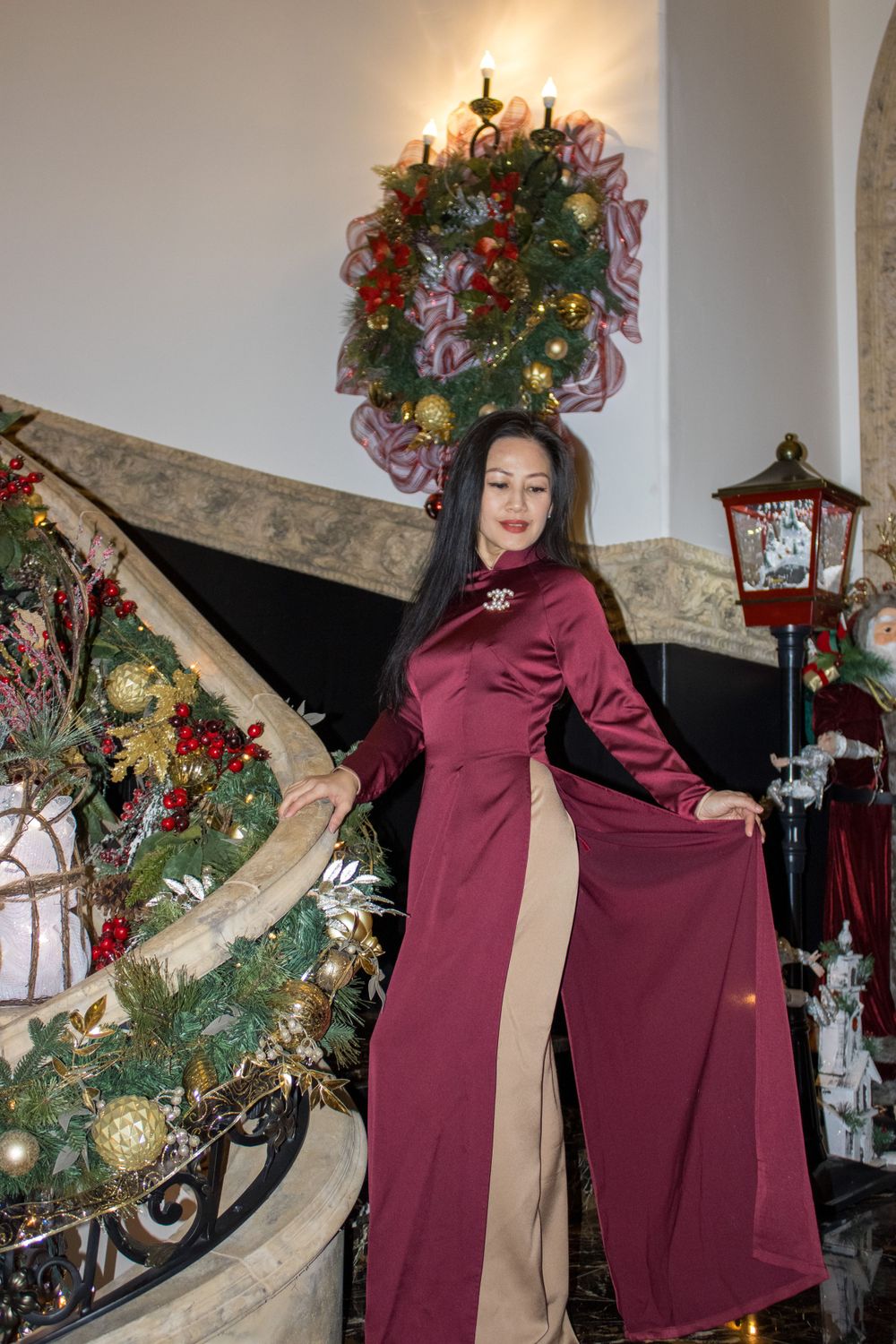 Woman in elegant burgundy ao dai dress poses near Christmas decorations on a staircase.