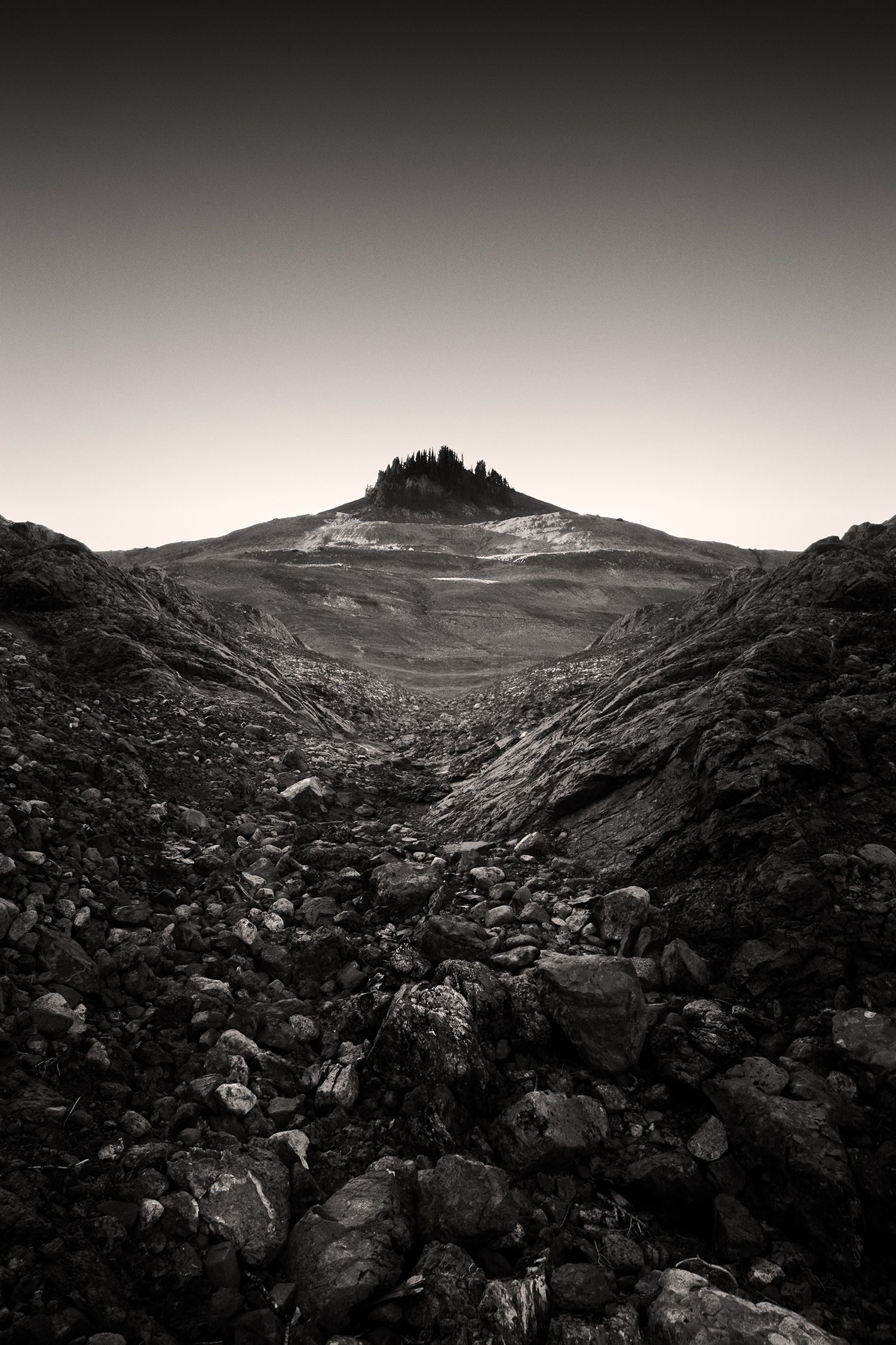 Black and white landscape of rocky valley leading to isolated hilltop peak.