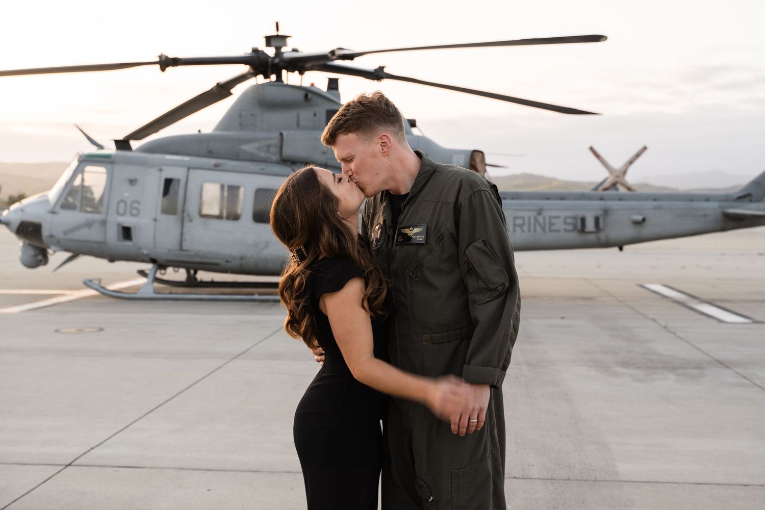 A military couple shares a romantic moment in front of a helicopter on a tarmac.