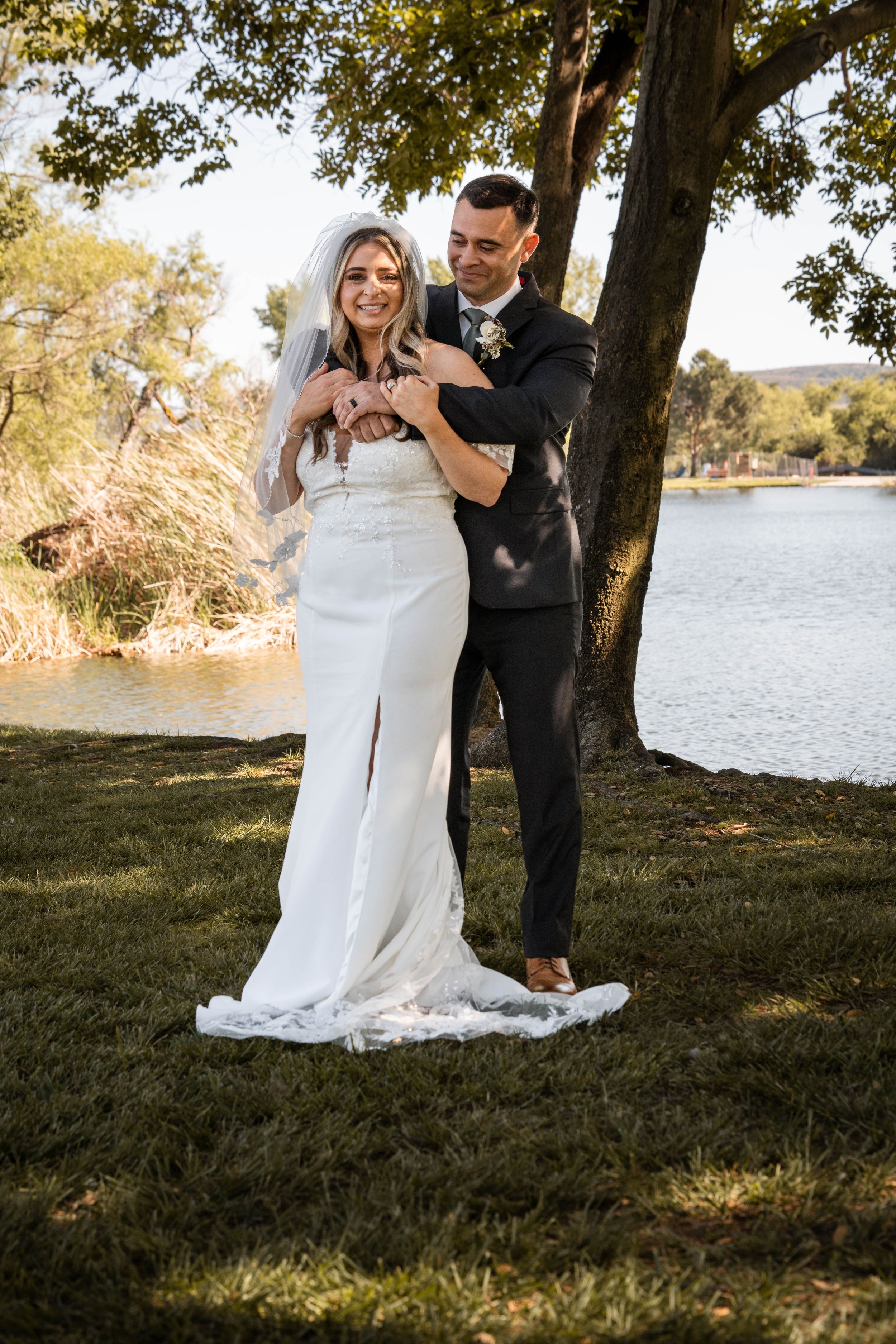 A romantic wedding photo showing a couple standing by a lake under a tree in golden sunlight.