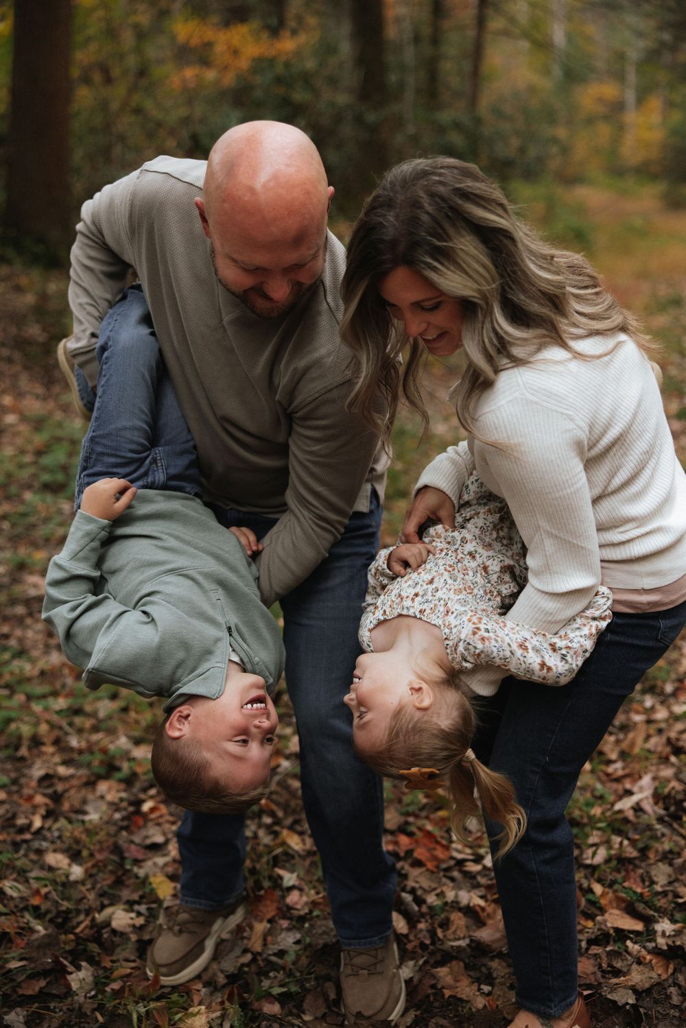 Laughing parents playfully flip their two children upside down in an autumn leaf-covered forest setting.