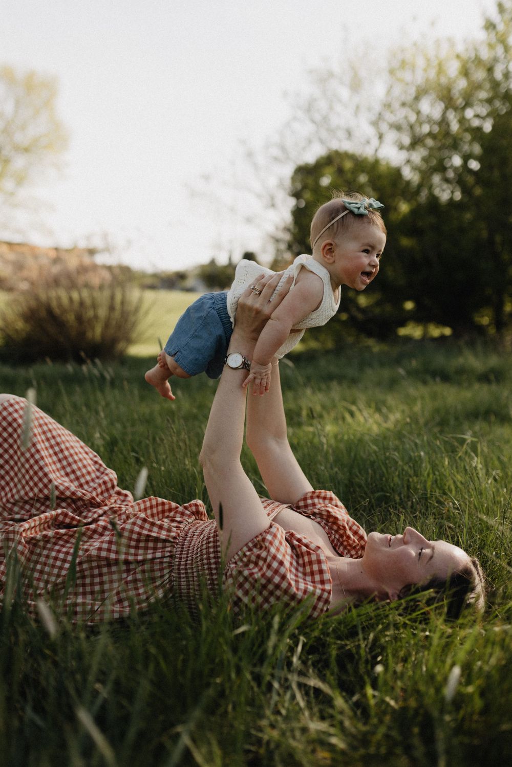 Mother lying in grass lifting smiling baby overhead during golden hour outdoor family photo session.