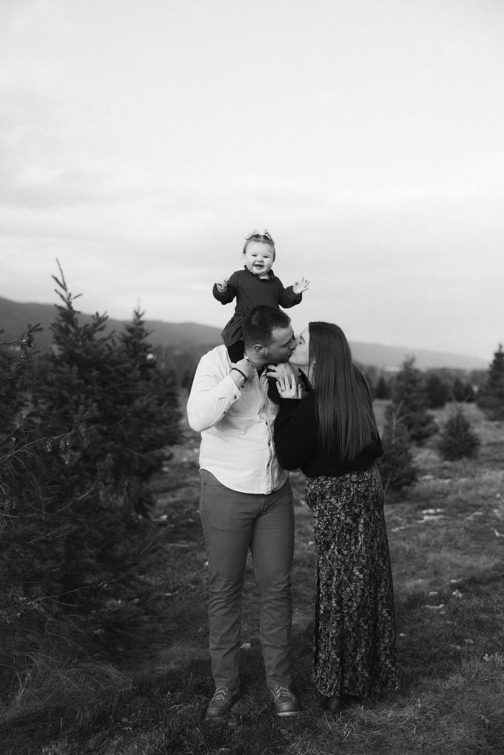 Black and white photo of a couple at a Christmas tree farm, with the man holding a laughing baby on his shoulders.