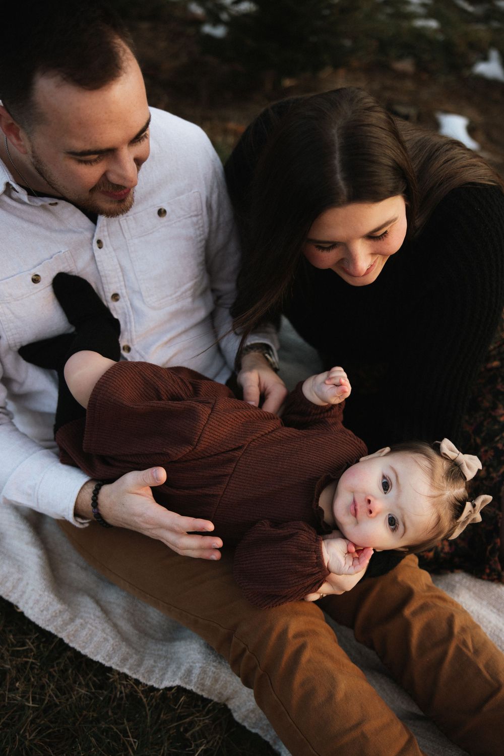 A young family sits on a blanket outdoors, parents smiling at their baby girl in a brown outfit amid a wintry backdrop.