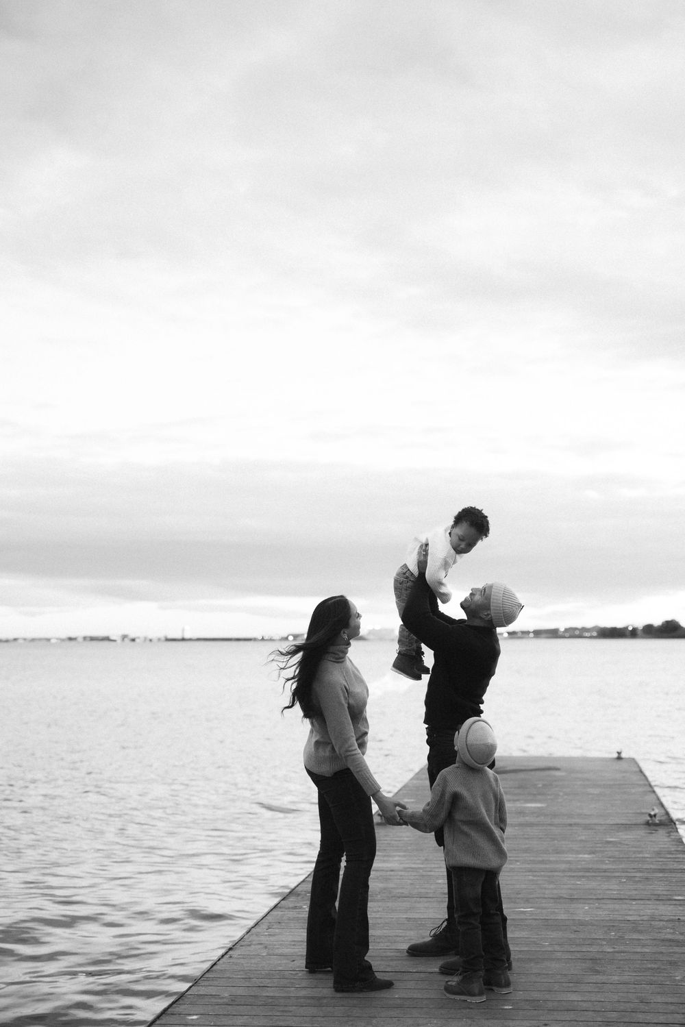 Black and white photo of a family on a dock, parents holding hands with child as dad lifts baby in the air.
