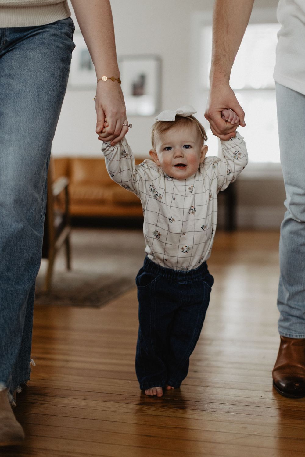 Baby taking first steps while holding both parents hands on hardwood floor at home.