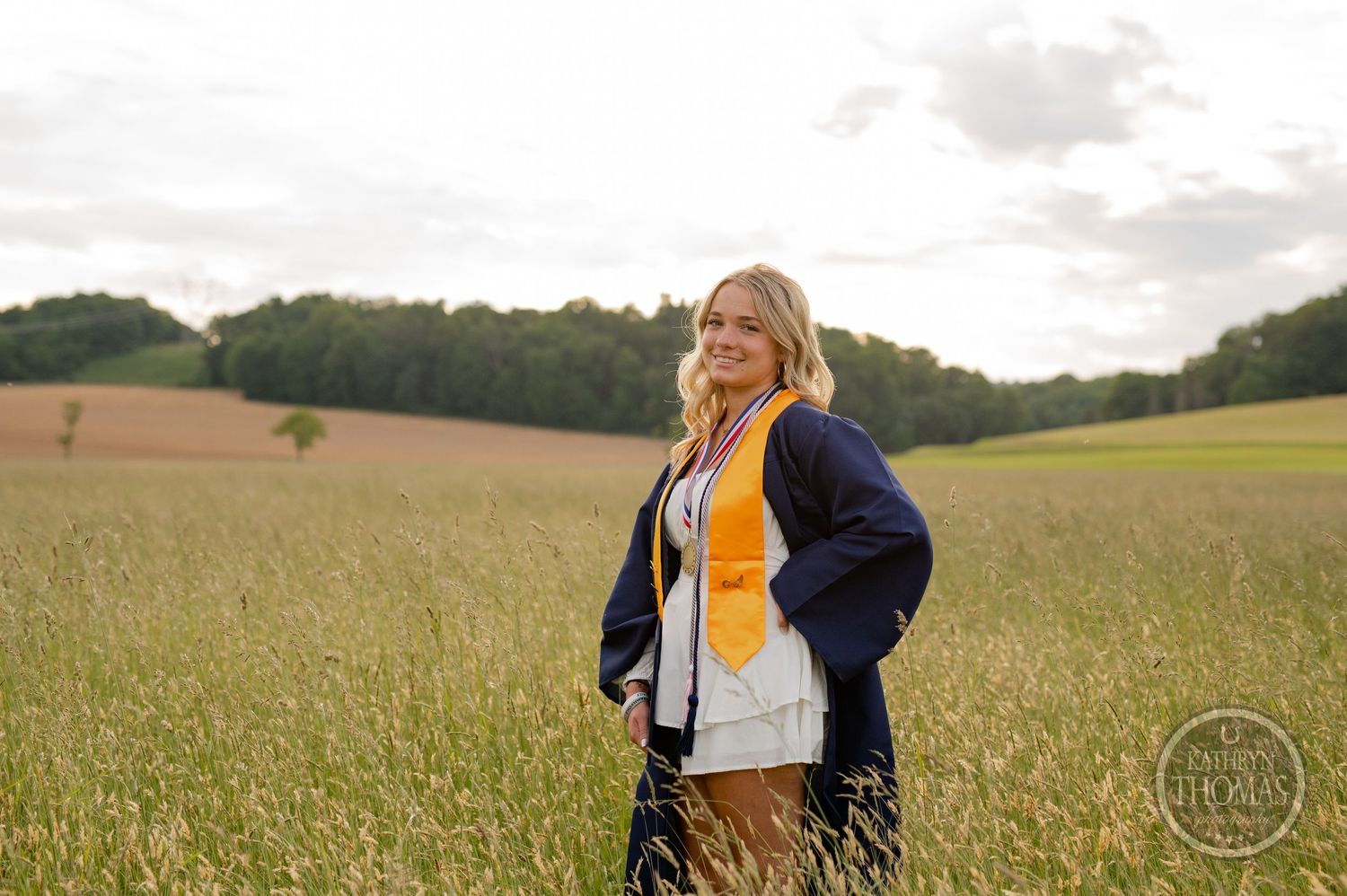 Graduate wearing navy and gold academic regalia poses in rural field at dusk.